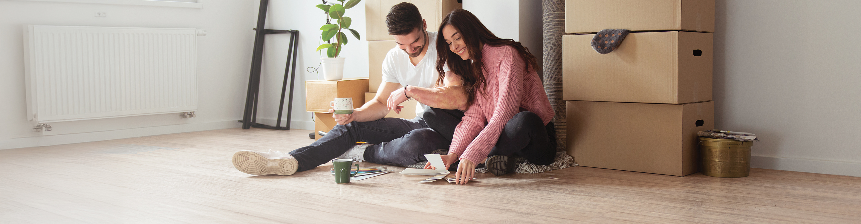 Couple sitting on new hardwood floors comparing flooring samples among moving boxes during the Top Floors Carpet One Simply Beautiful Flooring Sale in Suwanee